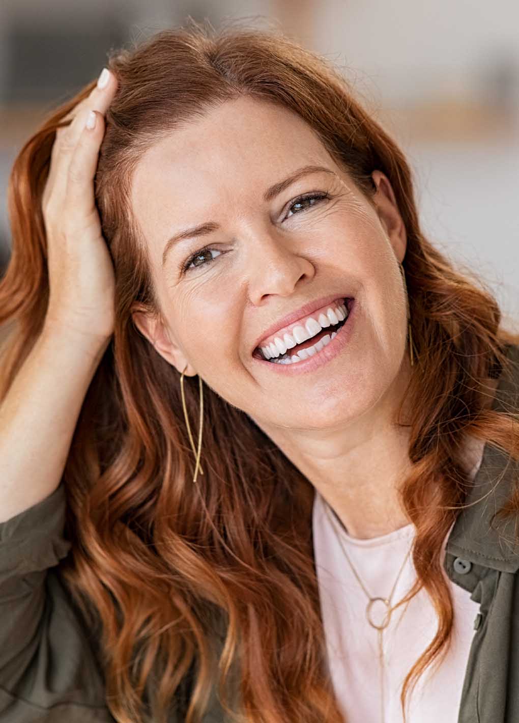Woman with red hair smiling and touching her hair, wearing a green jacket.