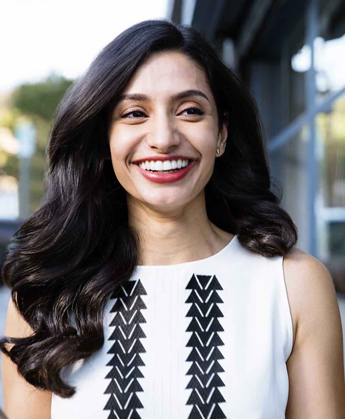 Woman with long dark hair wearing a white top with black patterns, smiling outdoors.