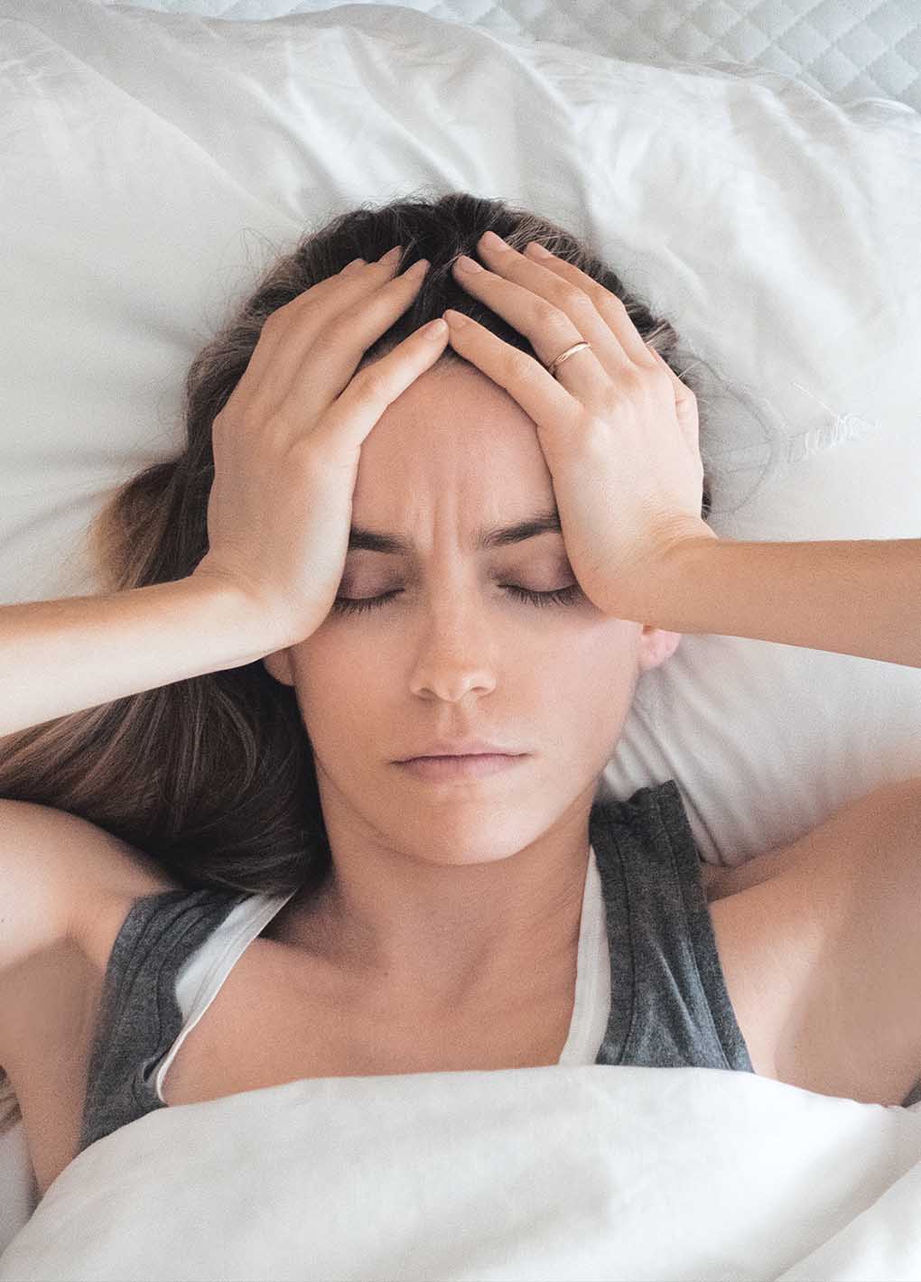 Woman lying in bed with hands on her head, possibly experiencing stress or discomfort.
