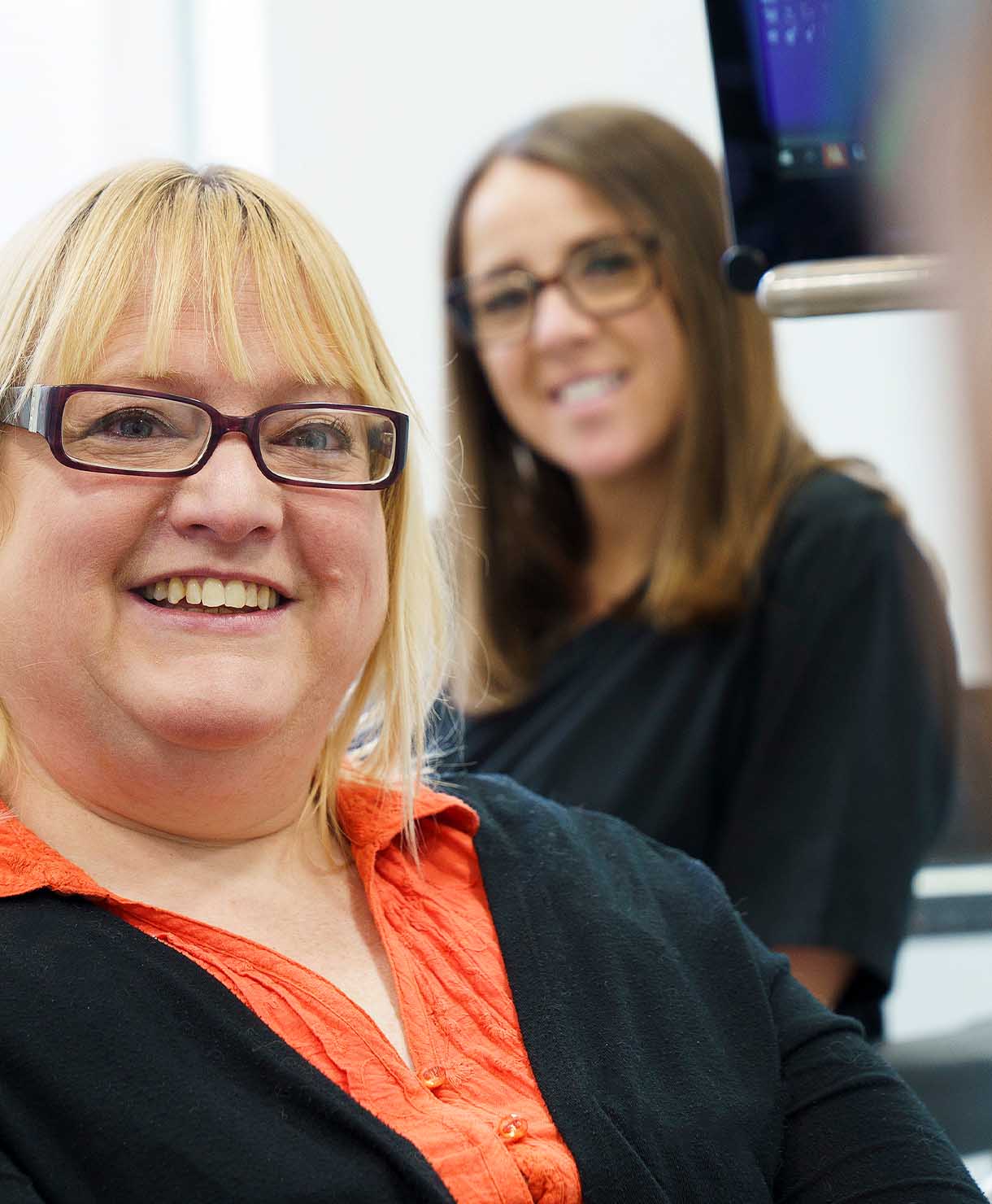 Two women smiling, one in the foreground with glasses and an orange shirt, the other in the background wearing glasses.