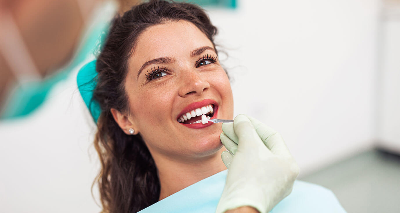 Brunette woman with red lipstick smiling in foreground looking up at staff member doing dental procedure with example tooth being held